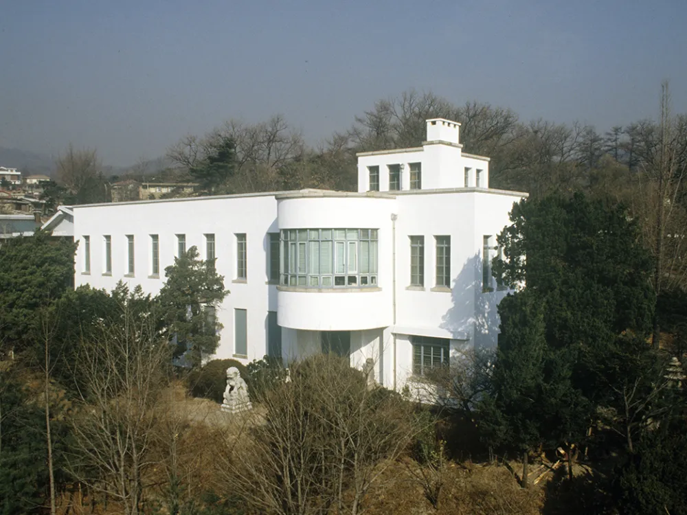 photo of a white building seen through trees with a stone lion sculpture visit near the center of the photo