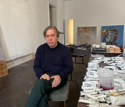 A man in a blue sweater sits in a chair in a large white room. To his left is a table covered in tubes of paint.