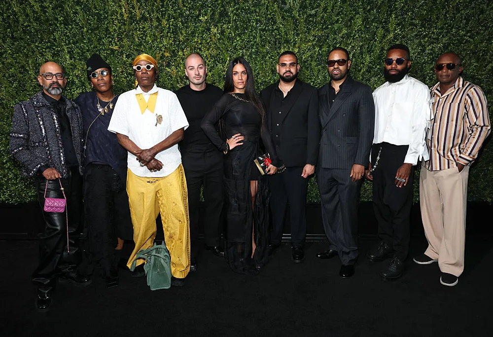 A group of nine stylishly dressed people standing side by side in front of a green faux plant wall