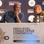 Two bald Black men sitting at a table with microphones, leaning towards each other in conversation