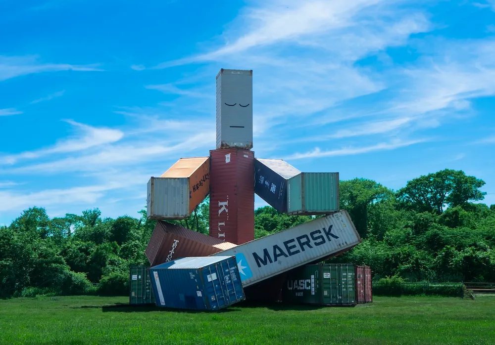 A towering sculpture by Matt Johnson made from stacked shipping containers forms a cross-legged meditating figure on a grassy field at The Ranch in Montauk, New York, beneath a bright blue sky.