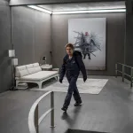 Willem Dafoe wears a jumpsuit and a backpack in a fancy apartment. On the wall is a picture of a person duct-taped to the wall.