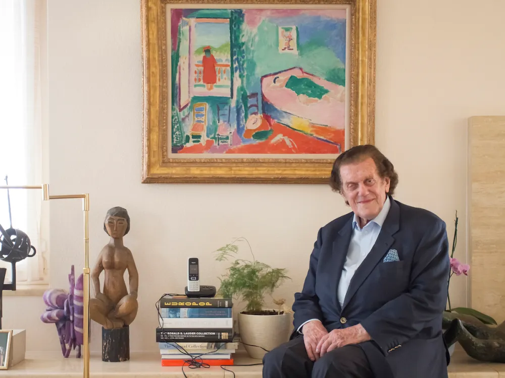 A man in a suit seated next to a stack of books and beneath a framed painting of a woman looking out over a balcony.