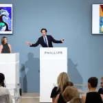 An auctioneer at a rostrum marked with the name of Phillips takes bids in a sale of a David Hockney painting of ocean waves that appears on screens behind him.