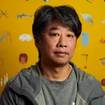 Portrait of Akira Ikezoe standing in front of a painting of a range of objects, including rabbits and a snail.