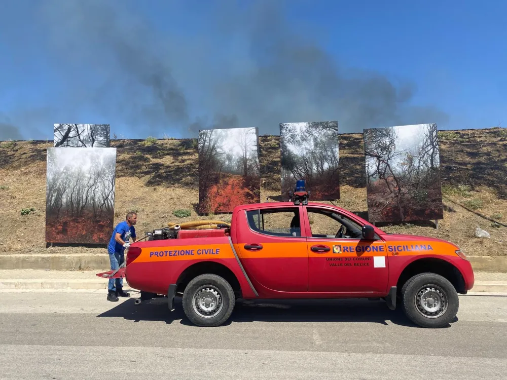A photography installation about wildfires by the artist Stefano Cerio in Gibellina, Sicily, was nearly destroyed by a real wildfire in early August, 2026. The image shows firefighters at the scene, where the ground around the installation is smoking and blackened from the extinguished flames, as smoke rises into the sky.