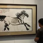 A man in a dark t-shirt stands in from of a ink on paper drawing of a galloping horse.