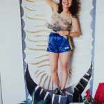 A color photograph shows a woman in running shorts holding paintbrushes in one hand and raising her other fist. Behind her is a mandorla outlining her body, and below her is a rug on which various objects are placed.