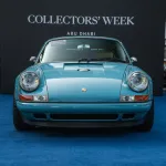 A light blue Porsche on a dark blue carpet surrounded by trees in the background.
