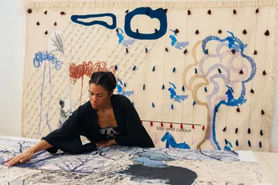 A woman holds down a textile piece on a workbench for an artwork in-progress with a finished piece behind her.