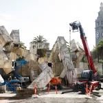 several construction cranes around a brutalist public artwork made of jumbled concrete tubes