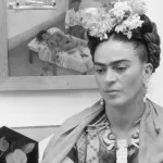 circa 1945: Mexican painter Frida Kahlo (1907 - 1954) sits with her arms folded, looking down, in front of one of her paintings and a wooden bird cage. She wears flowers in her hair and a wooden necklace. (Photo by Hulton Archive/Getty Images)