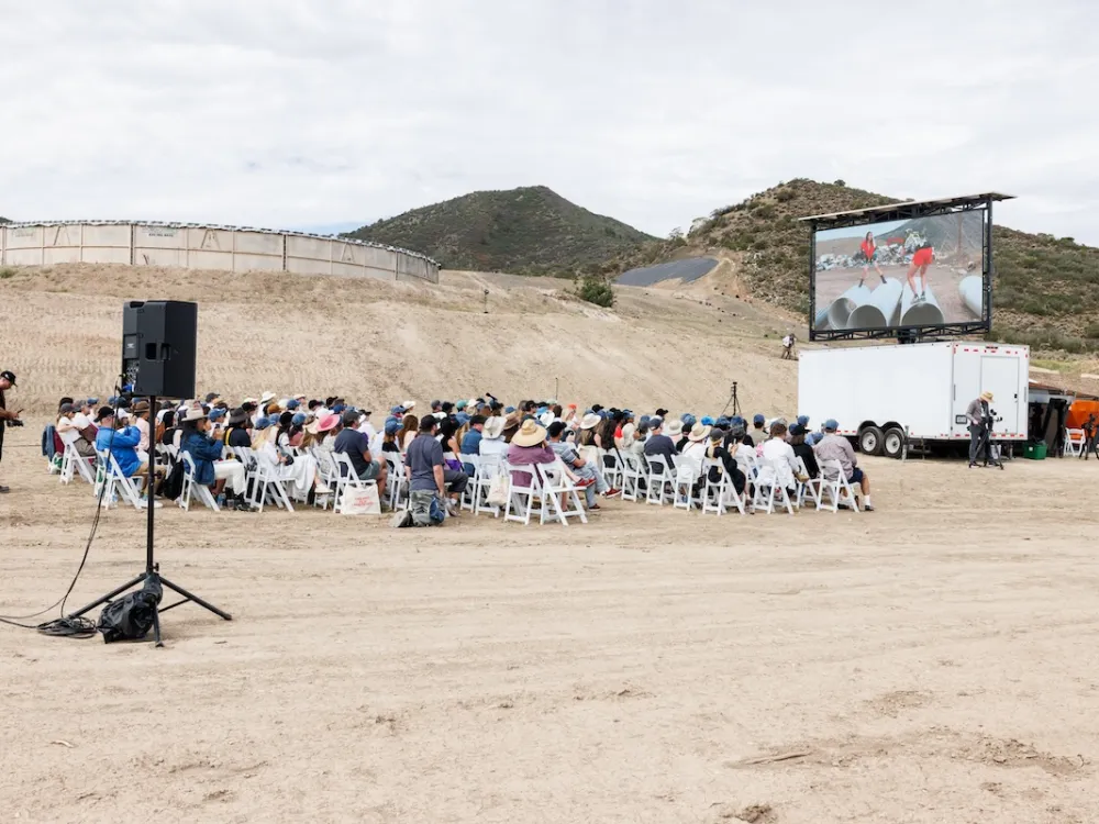 An audience seated in a barren outdoor landscape in front of a large screen.