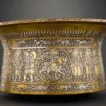 A gold-colored metal vessel with figures carved into its sides.