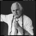 A black-and-white photo shows a white-haired, mustachioed white man with long ponytails, artist Milan Knížák, posing for the camera in a white shirt and tie.