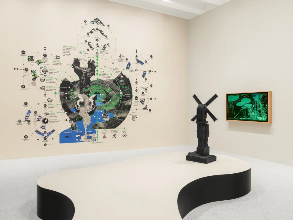 A photo shows an art gallery with works on display. A large wall-printed display resembles a legend, and a green-hued video is displayed on a monitor on the wall. On a biomorphic platform at the gallery
