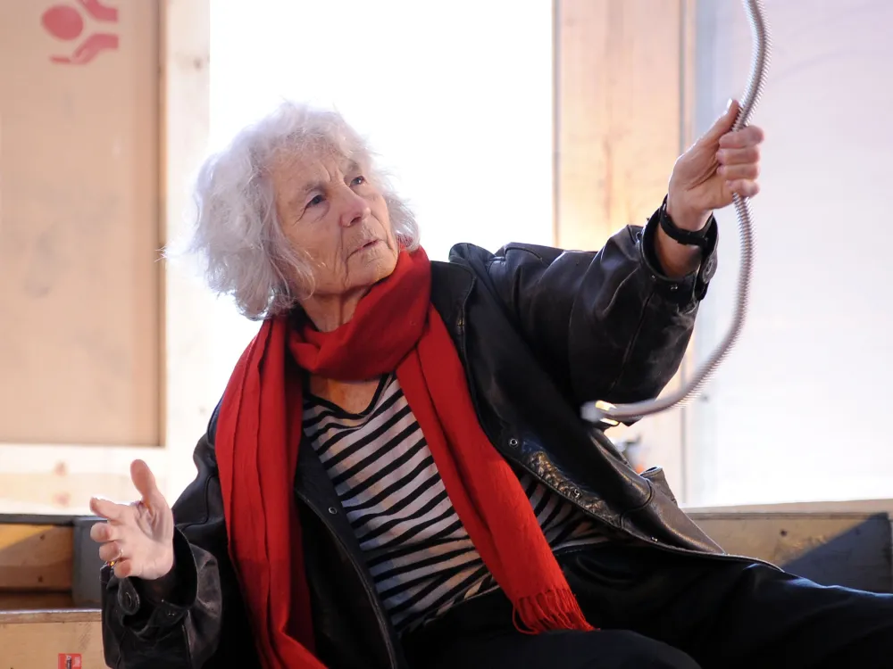 an older woman with white hair, and black and white striped shirt, and a red scarf, seated and leaning in an art studio while holding on to a rope