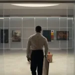 A man seen from behind holds a painting as he looks at a storage shelf holding four paintings from different eras.