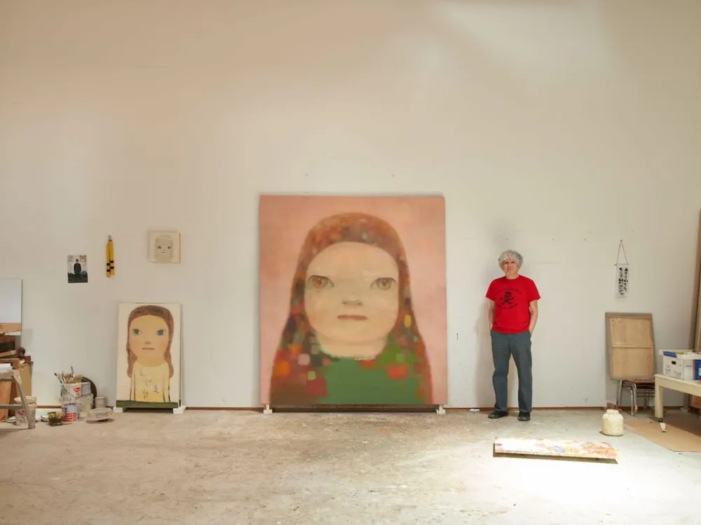 A man standing in his studio with paintings of faces against the walls.