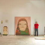 A man standing in his studio with paintings of faces against the walls.