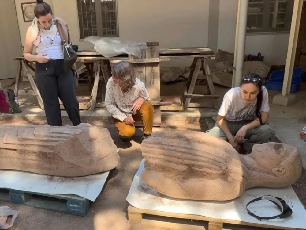 Four people surround a seven-foot-tall marble sculpture of a man, lying on its back on wooden pallets on the ground