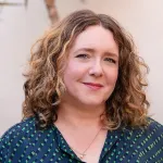 Woman with curly reddish-brown hair smiling in a navy patterned blouse against a beige background.