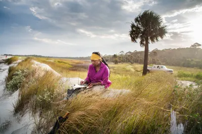 Suzanne Jackson sits on a grassy beach on a barrier island as she draws. The lighting is dramatic