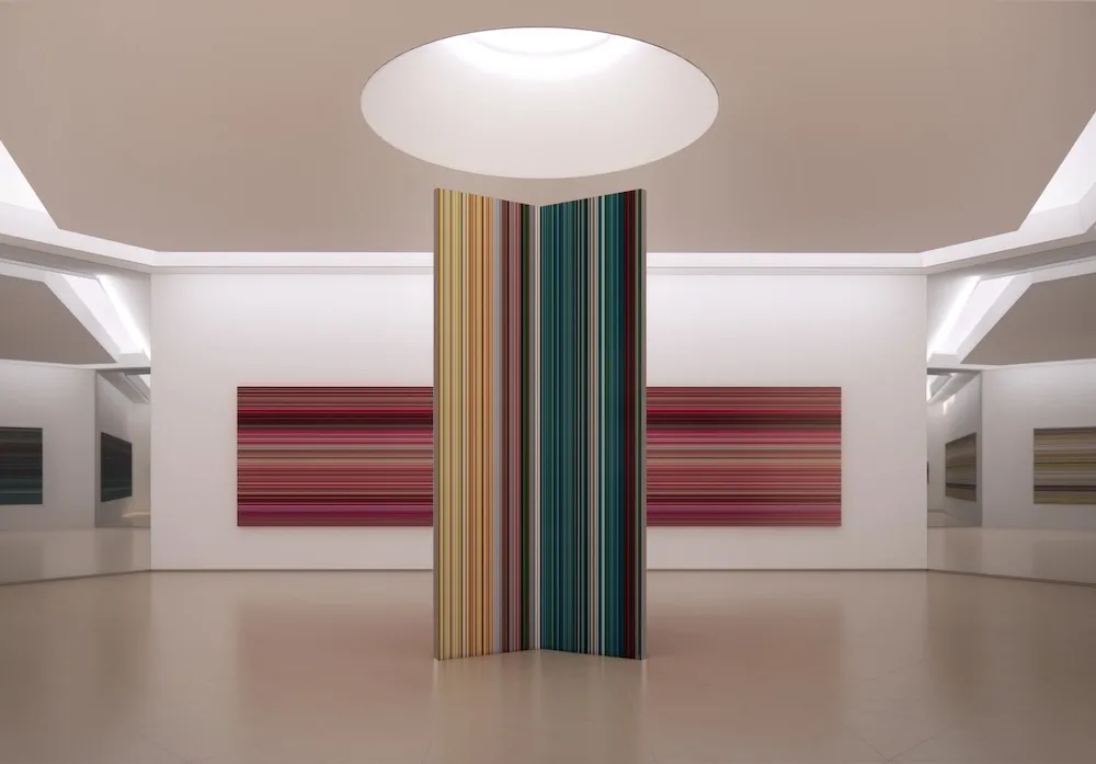 A white room bearing large colorfully striped paintings, with a skylight at top.