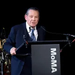 A man in a dark blue suit, white shirt, and tie standing on a stage behind a lectern with