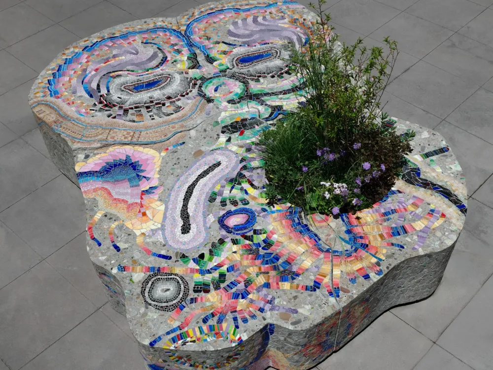 overhead view of an oblong stone bench covered in a colorful mosaic pattern with a plant growing through it