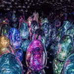 Mirrored room filled with glowing, egg-shaped sculptures in jewel-toned blues, pinks, purples, and greens, with a visitor standing among them.