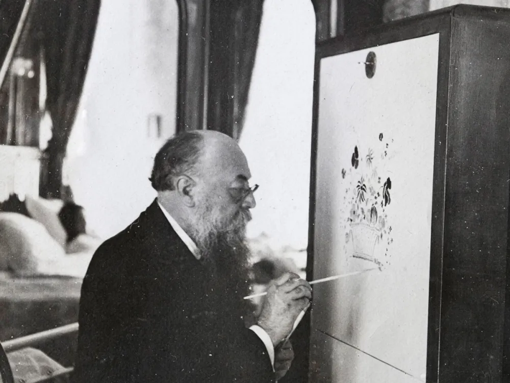 Archival photograph of a man with a long beard in a suit painting a floral scene on a piece of furniture.