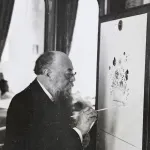 Archival photograph of a man with a long beard in a suit painting a floral scene on a piece of furniture.