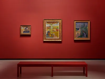 View of three Christ-themed Gauguin paintings hanging on a red wall. 