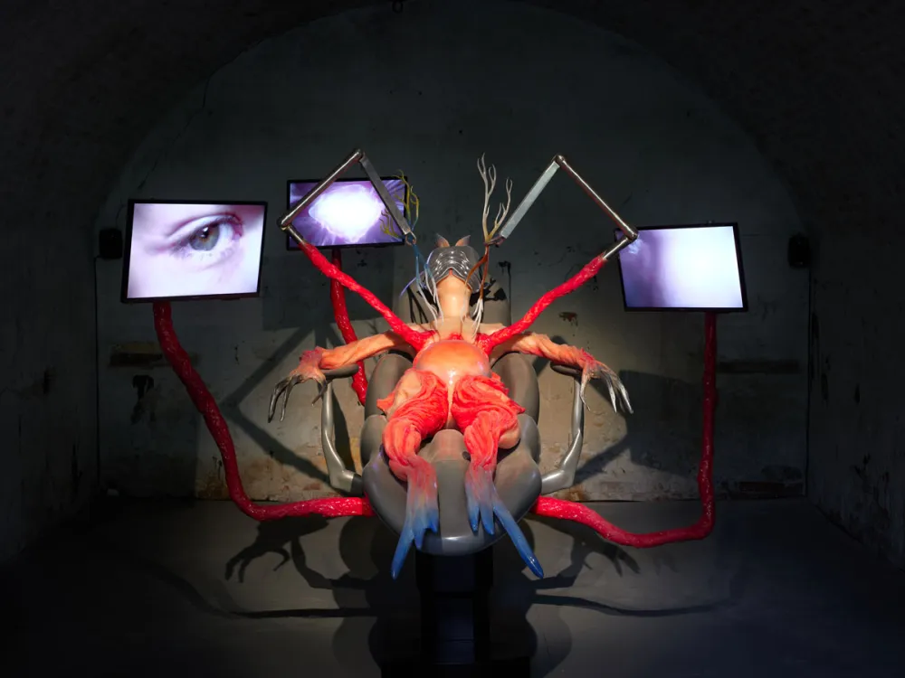 View of a sculpture of a humanoid figure who appears to be flayed surrounded by three monitors that play videos.