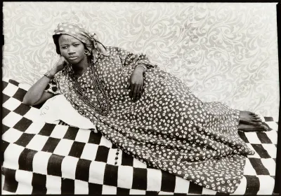A black-and-white photograph of a Black woman in a floral dress laying like an odalisque on a checkered blanket.