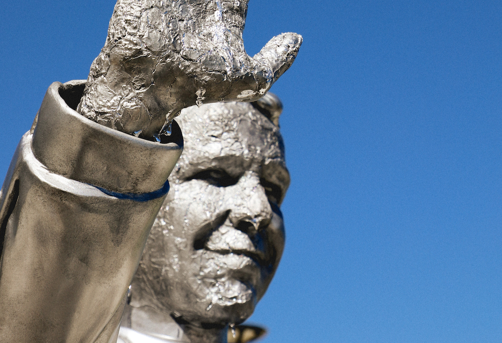 Close-up of a silver-colored statue of Ron DeSantis with its right hand raised, its face and hand beginning to melt against a blue sky.