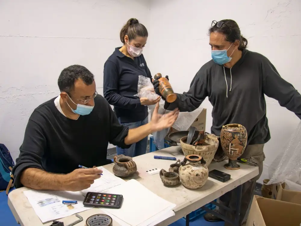 three people wearing facemasks examining ancient vessels on a table