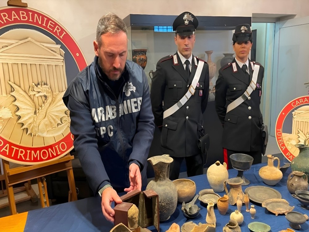 Italian Carabinieri art-crime officers stand behind a table displaying recovered ancient artifacts—terracotta vases, jugs, bowls, and oil lamps—during a press presentation, flanked by two large Carabinieri