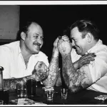 A vintage black-and-white photo of two tatooed men arm wrestling and smiling.