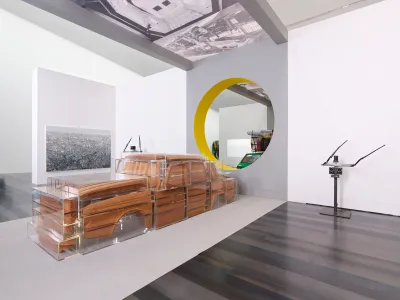 View of a museum exhibition showing a sculpture of a wooden car that has been cut up into pieces and encased in various plexiglass containers.