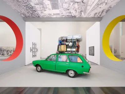 View of a green car that is overload inside and has more stuff on top in a museum between two circular openings.