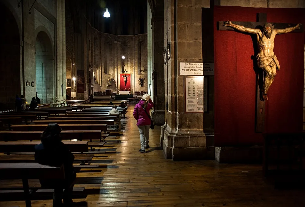 Interior of a Spanish church with rows of wooden pews and a large crucifix displayed against a red backdrop.