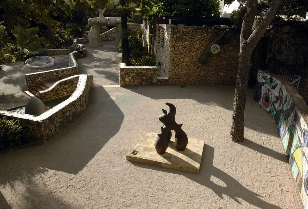 Sunlit sculpture garden at the Fondation Maeght in southern France, featuring abstract sculptures, stone walls, and a colorful mosaic mural.