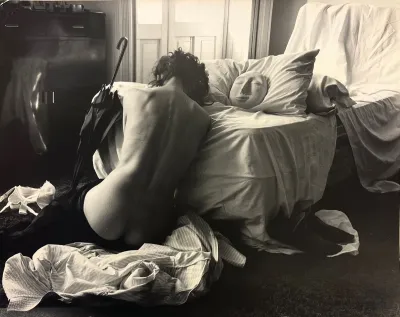 A vintage gelatin silver photograph showing the back of woman (who is shirtless and pants are pushed down) leaning on a bed. Next to her on a pillow is a mask.