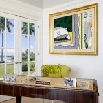 A photograph shows the white-walled interior of a room in a home, with palm trees, grass and water visible through the windows. Over a desk hangs a Pop art painting by Roy Lichtenstein showing a wall with parts of two paintings on display, one showing a sculpture of a head resting horizontally on a tabletop.