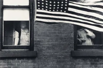 A photograph of a US flag waving in a front of a brick building with figures whose faces aren